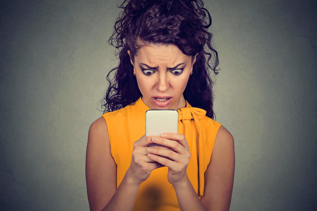 A frowning, anxious woman looks at her phone in shock and horror.