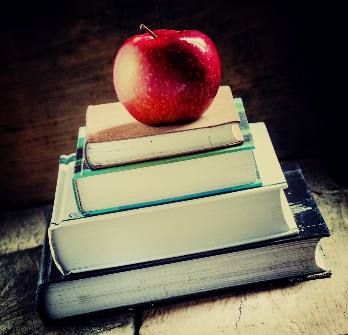 A stack of school books sits on a desk, with an apple on top. [Education/Learning]