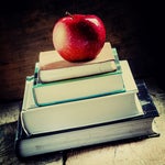 A stack of school books sits on a desk, with an apple on top. [Education/Learning]