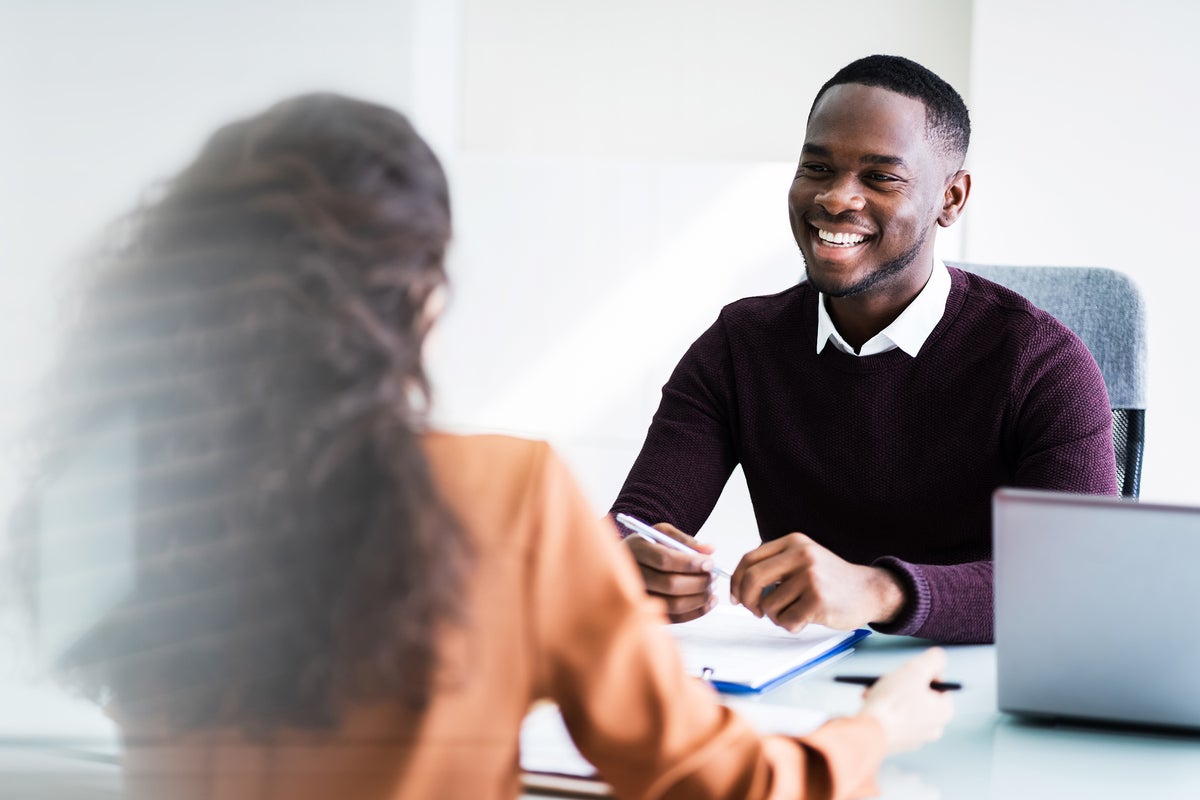 A man and woman sit on opposite sides of an office desk, in discussion.