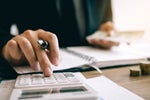 One hand uses a calculator while the other holds banknotes.