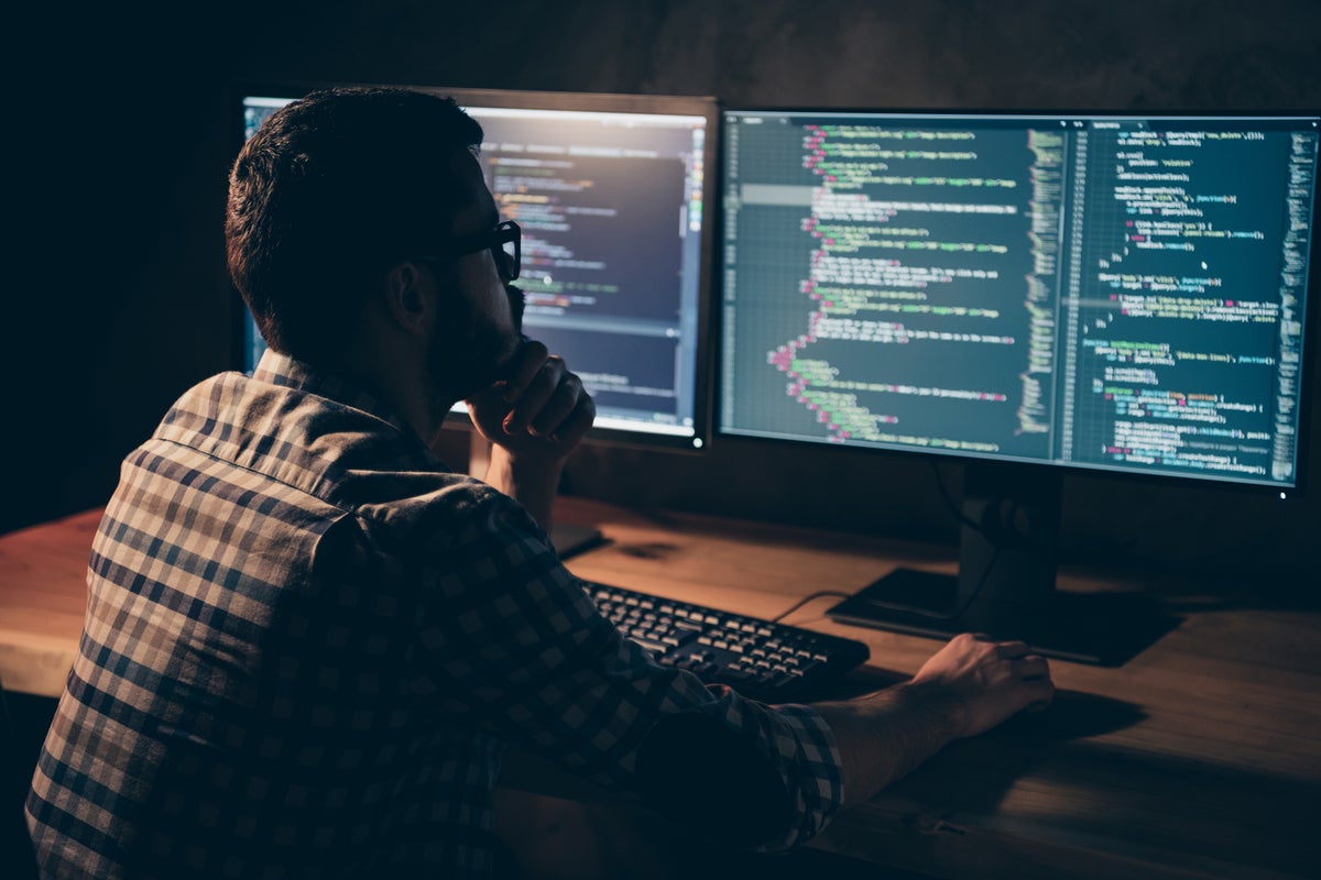 A developer works across multiple displays showing lines of code in a dimly lit workspace.