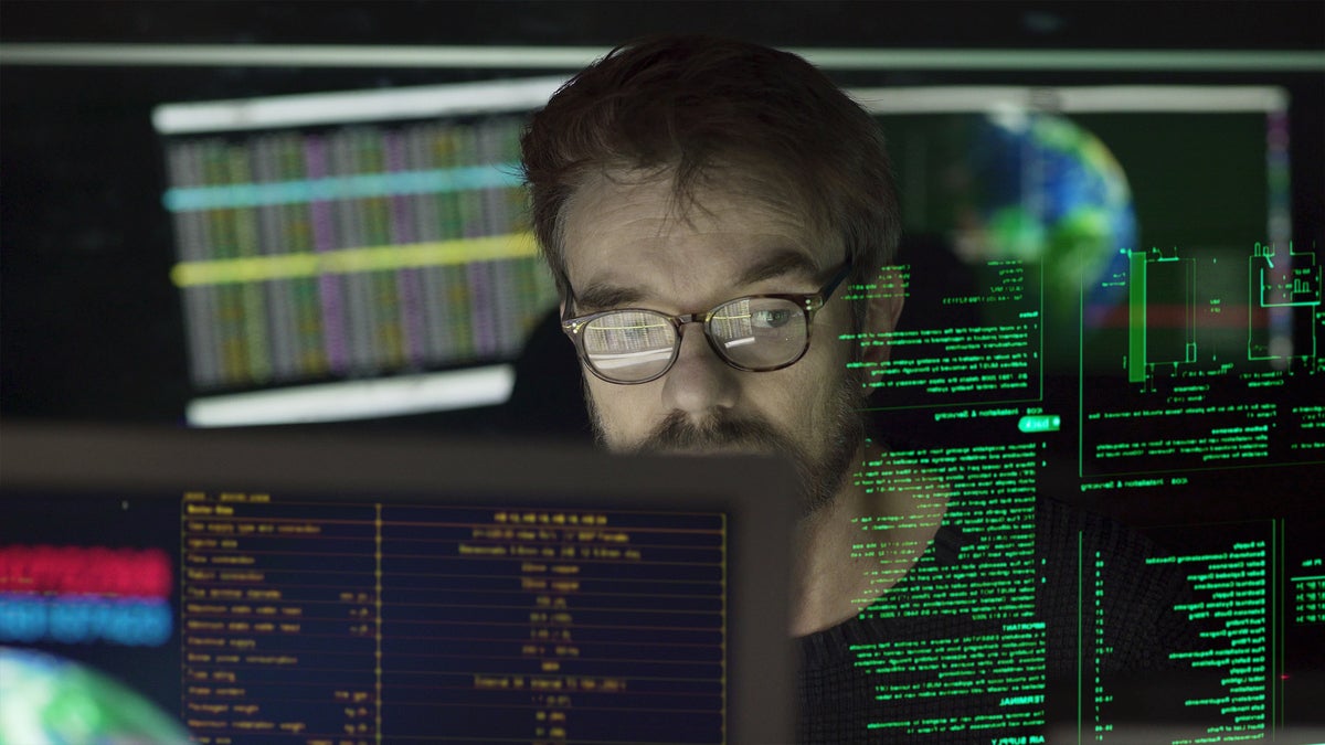 mature man surrounded by monitors & a holographic display which he is reading.