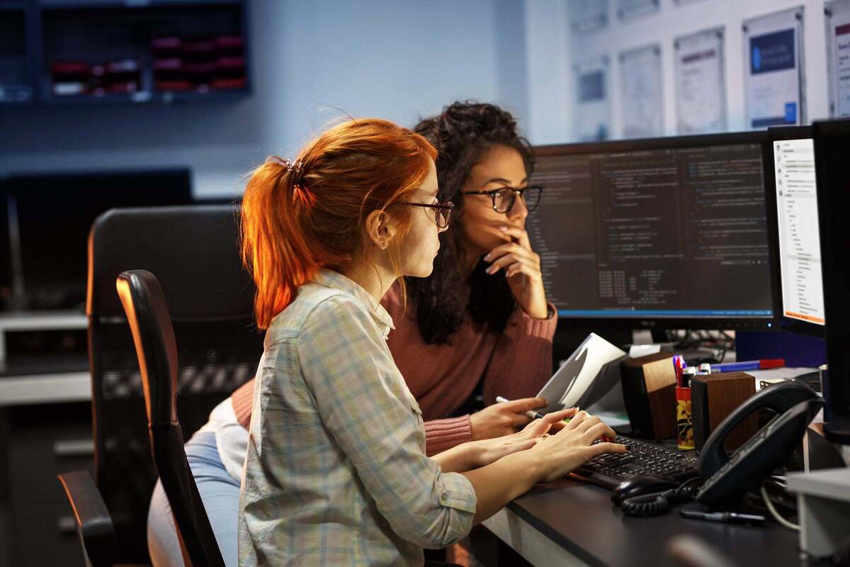 Two developers collaborate on a project as they review code on a display in their workspace.